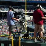 Two men experiencing fan interactions with a television set at a baseball game.